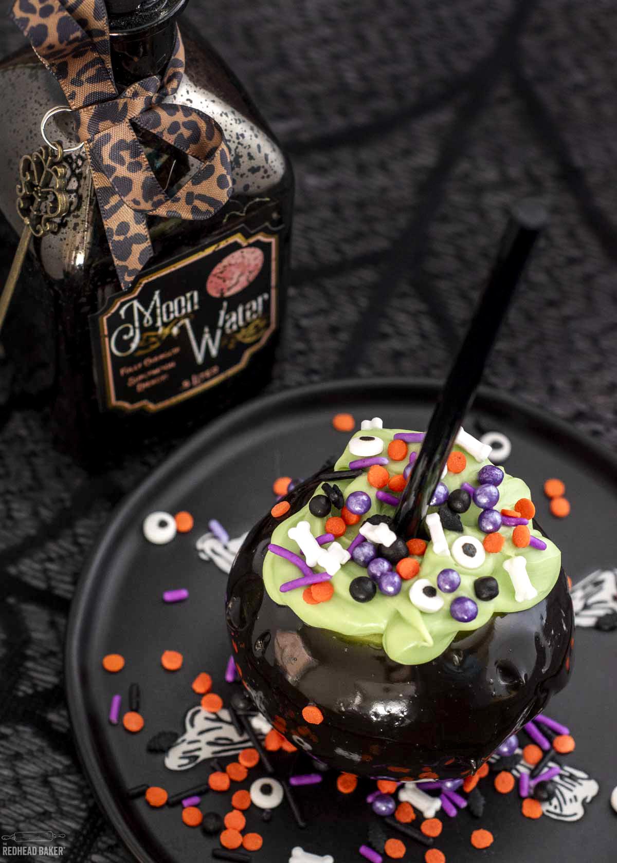 A cauldron caramel apple on a black plate in front of a bottle labeled "Moon Water".