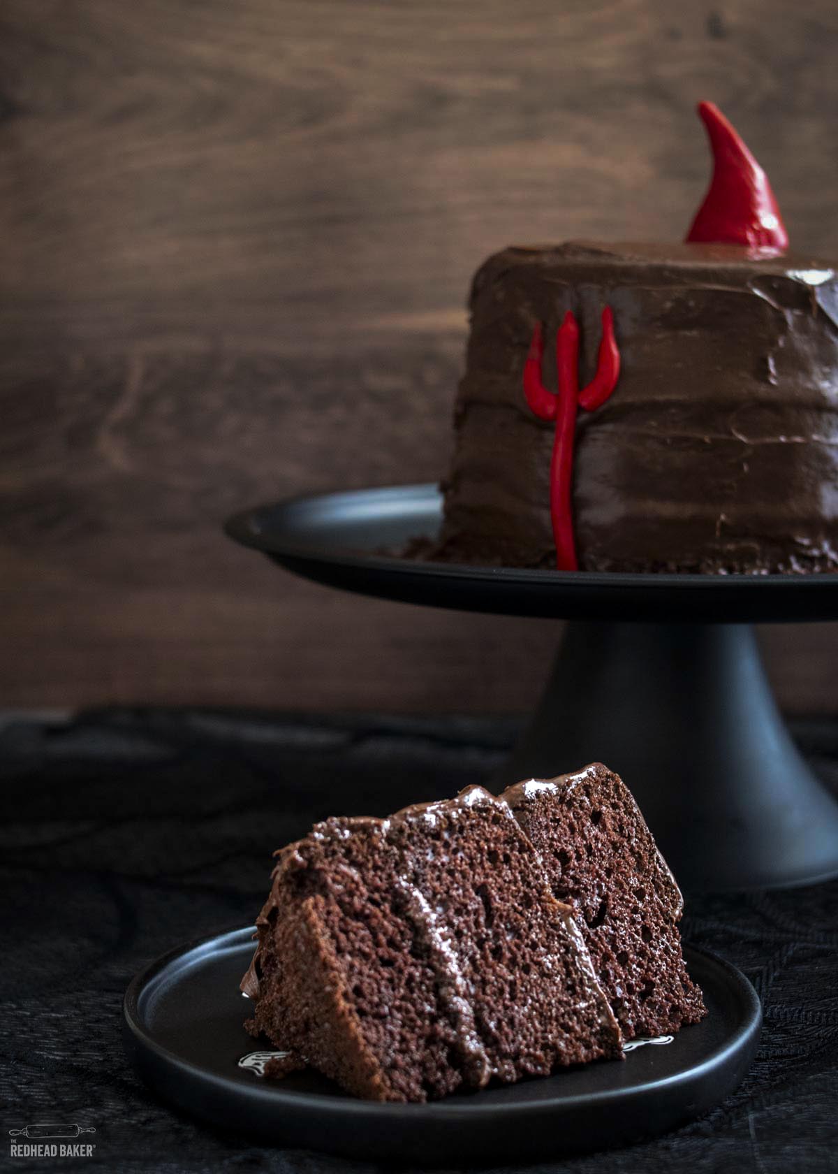 A slice of devil's food cake on a black plate with the rest of the cake in the background.