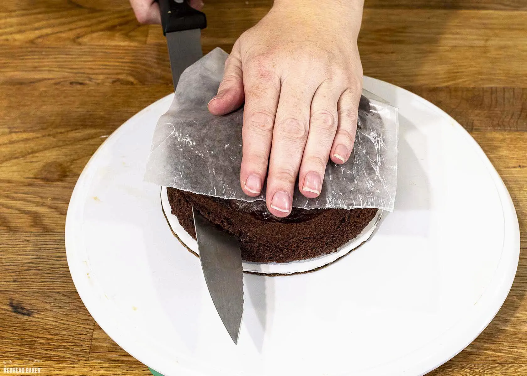 A hand on top of a layer of cake with a serrated knife demonstrating how to level a cake.