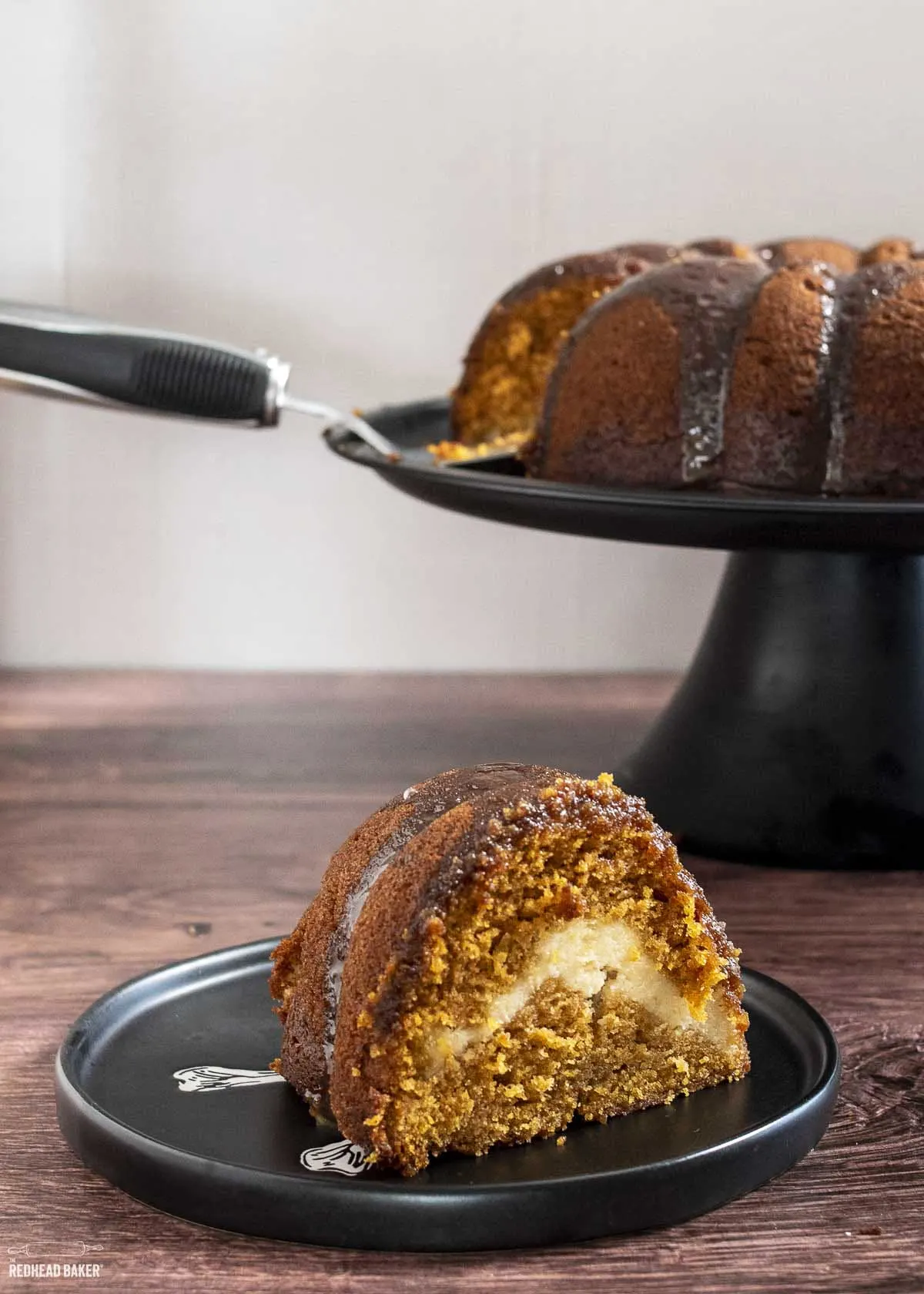 A slice of pumpkin bundt cake on a plate in front of the cake stand. 