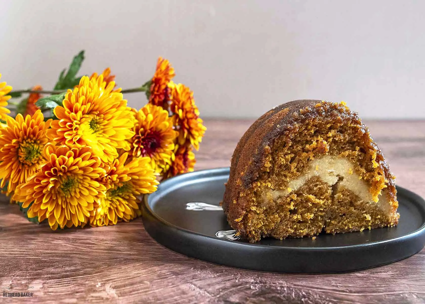 A slice of pumpkin cake on a black plate in front of a spray of fall flowers. 