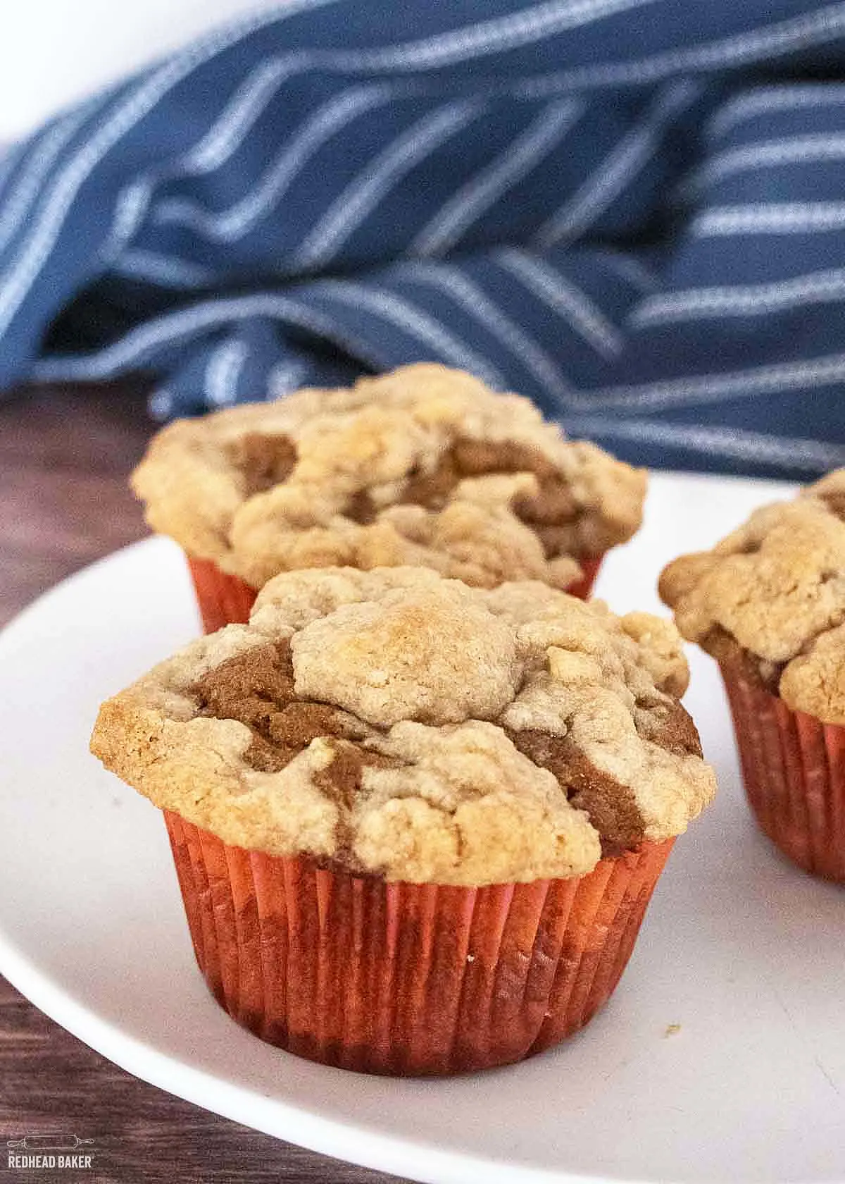 Three pumpkin muffins on a white plate in front of a blue tea towel. 