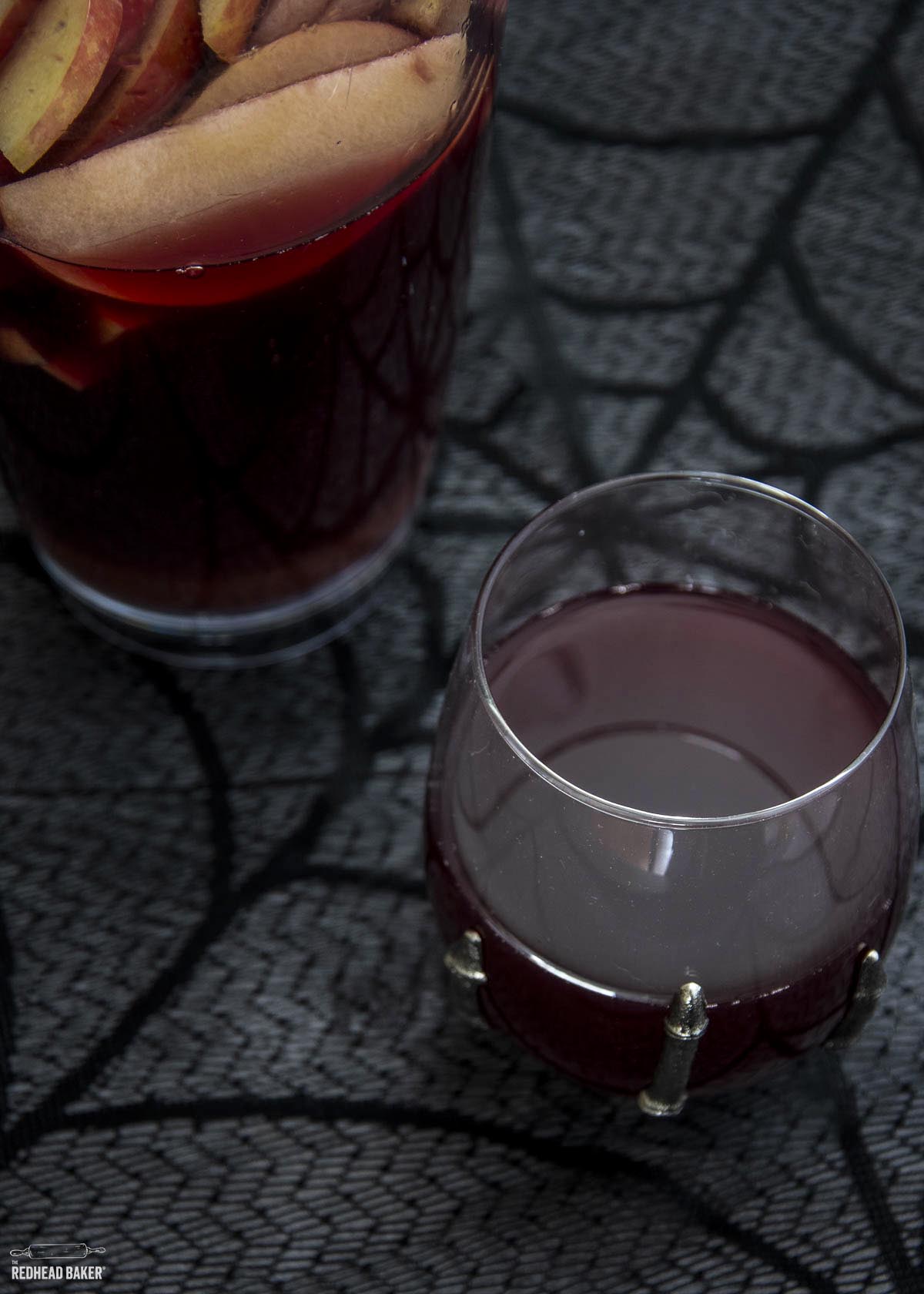 A glass of red cider sangria in front of a pitcher of more sangria. 