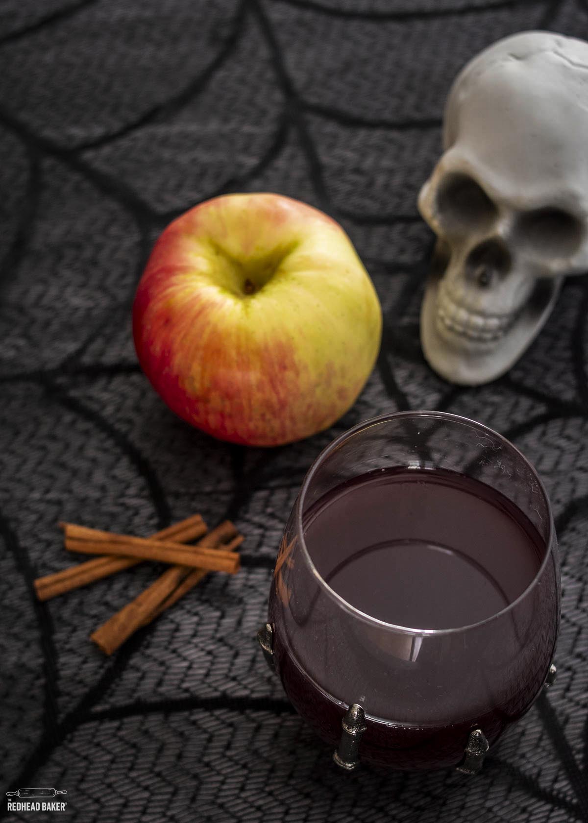 A glass of sangria next to three cinnamon sticks, a honeycrisp apple and a plastic skull decoration.
